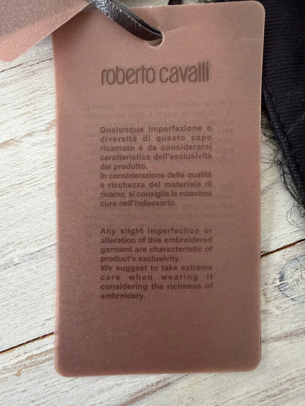A close-up of a brown garment tag with embossed text reading Roberto Cavalli, placed on a light wood surface.